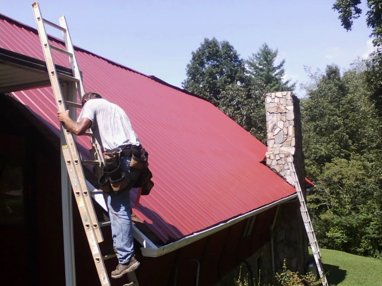 Roofer on a ladder beside a red metal roof and stone chimney — Soffit & Fascia Repair by Roofing Brothers in Berry Creek
